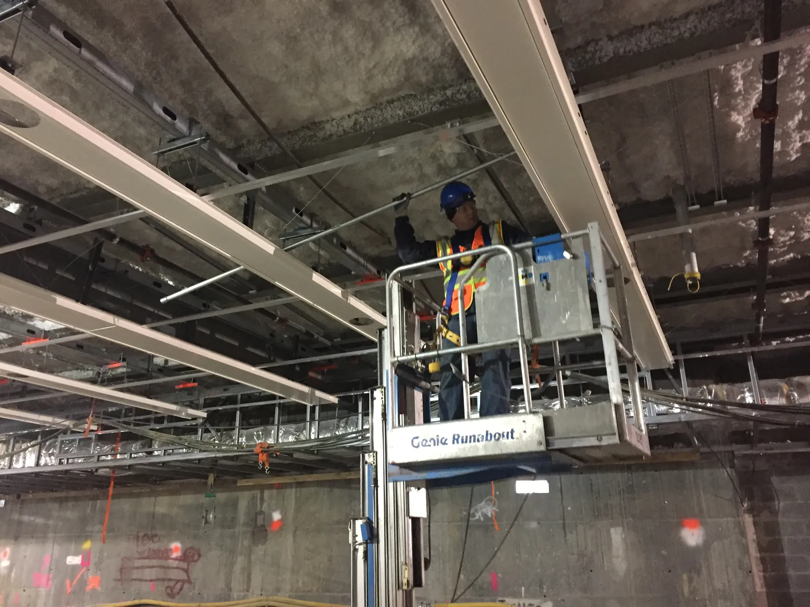 An electrician on a lift hangs lighting conduit in the ceiling of a large passenger concourse under construction