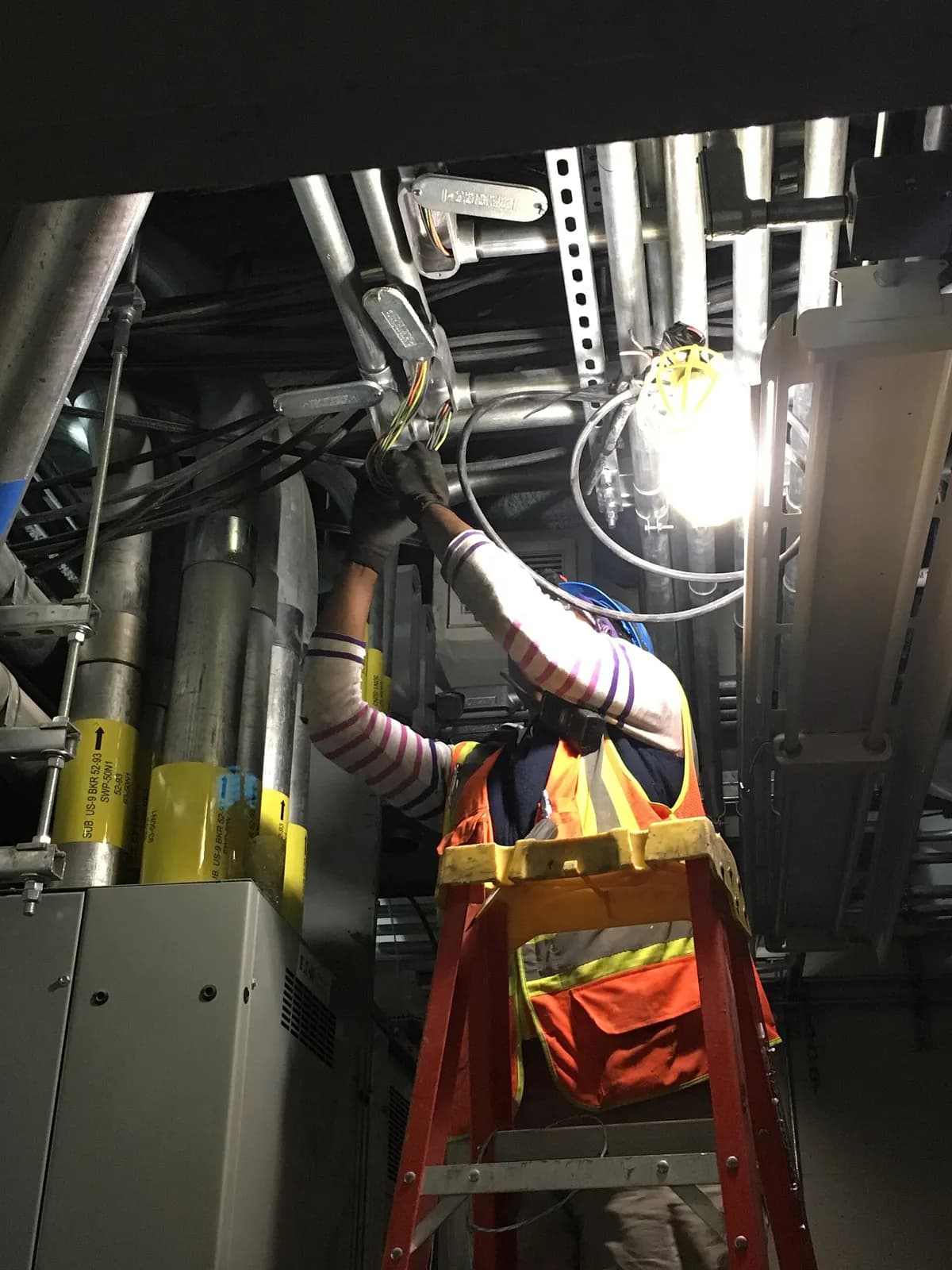 An electrician on a ladder installs conduit overhead inside an MTA ventilation facility in New York