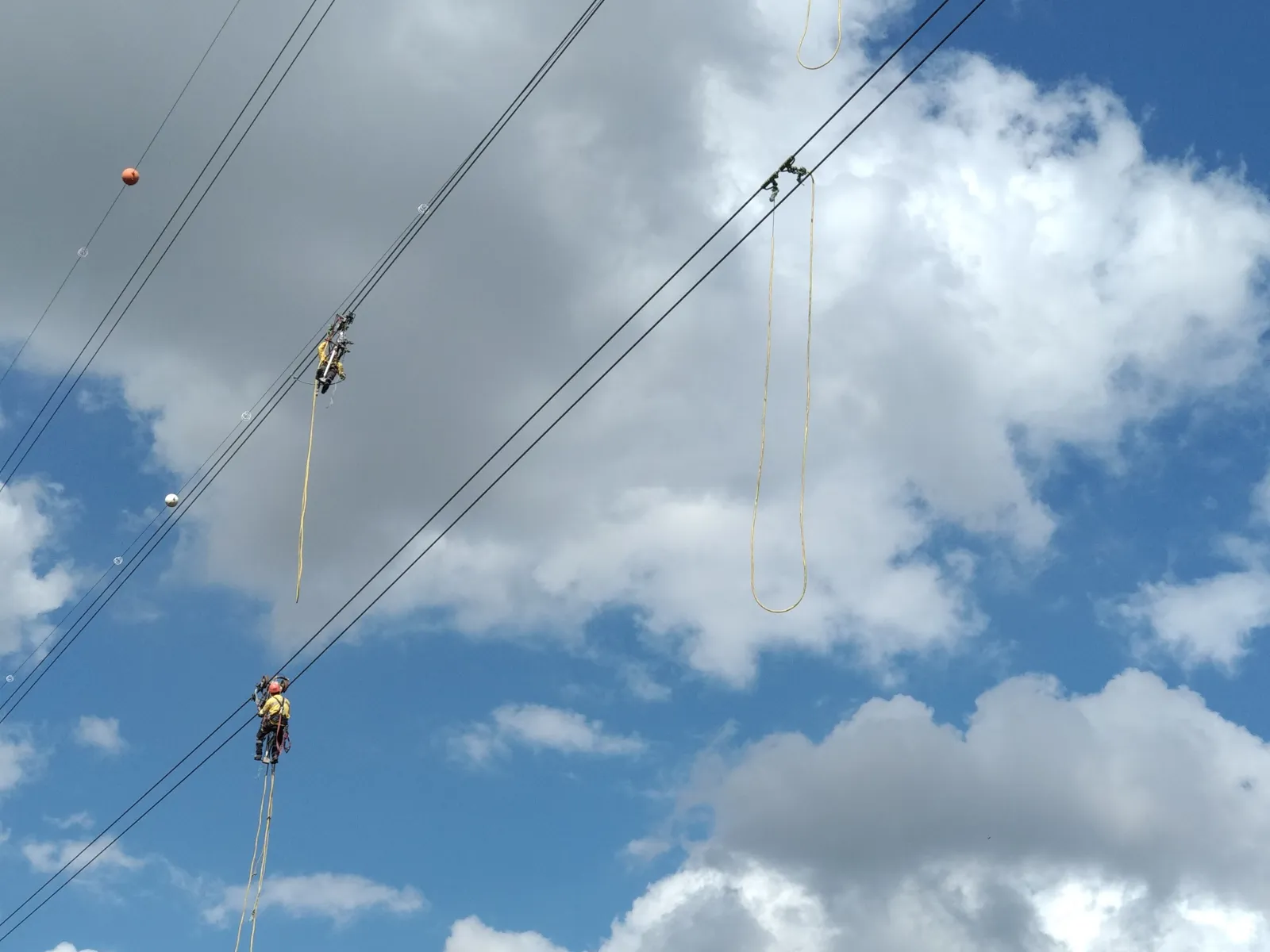 Line workers maintain an overhead high-voltage power line against a cloudy sky